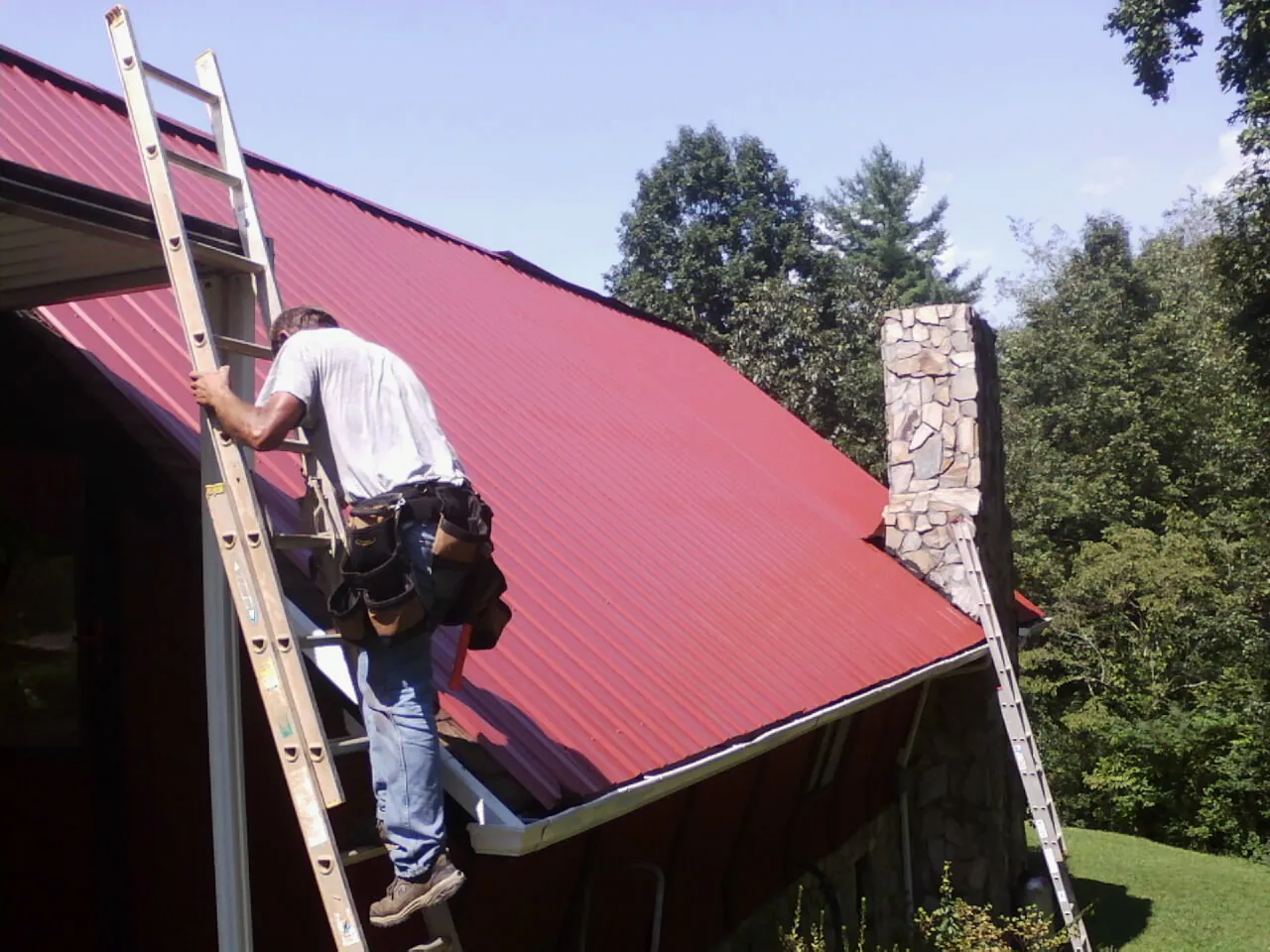 Roofer on a ladder beside a red metal roof and stone chimney — Soffit & Fascia Repair by Roofing Brothers in Kimberly